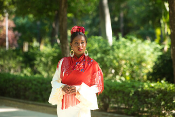 Young black woman dressed as a flamenco dancer stands in a famous park next to a duck pond in Seville, Spain. She wears a beige dress with ruffles and red shawl and flowers in her hair.