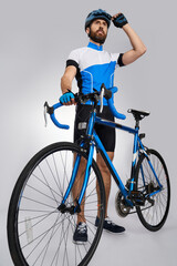 Strong motivated male cyclist getting ready to start in studio. Low angle view of bearded biker with blue bike looking away, while wearing helmet, isolated on grey. Concept of cycling, sport activity.