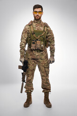 Serious male soldier with rifle in hand, posing in studio. Front view of bearded military man in camouflage gear, holding shotgun, looking camera, with gray background. Concept of military force.
