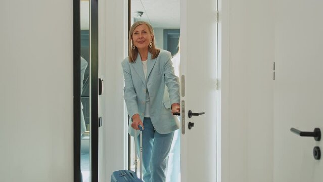 Two charming women elegantly dressed entering in modern hotel room. Smiling ginger businesswoman with mother rolling suitcases and looking at beautiful modern gorgeous interior indoors