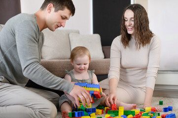 Happy family: dad, mom and child play with a toy constructor, sit on the floor at home, have fun together on the weekend