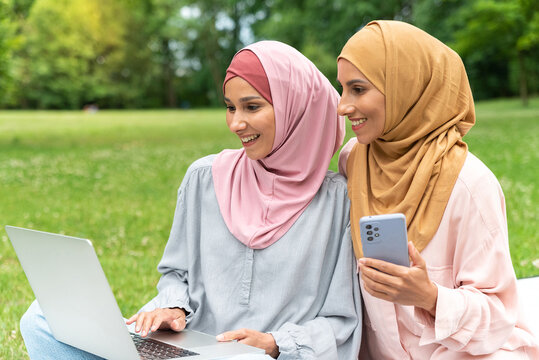 Portrait of two young happy Muslim women in hijabs look at computer screen typing use laptop and phone for shopping or work in a park. Girls browsing social networks have good news and exam results