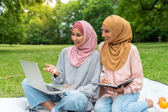 Two cute Muslim women sitting in nature in a city park listening using a laptop to a webinar ask questions write down comments. Girls study with a tutor and prepare for exams. Pleasant video call