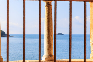 View through a barred window of a weathered column in front of the sea in the town of Rovinj in Croatia on a summer day with blue sky