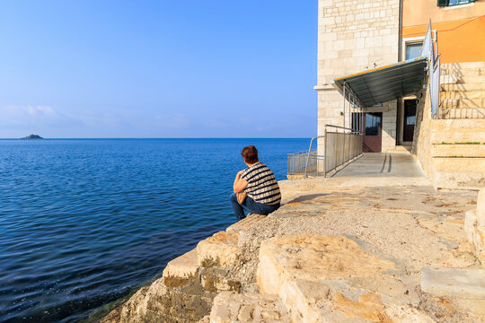 Elderly Caucasian Woman With Short Hair From Behind Sits On Steps Of Harbor Wall Of Croatian Town Rovinj And Looks Out To Sea To Small Island