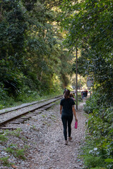 Woman walking backwards beside the train track on the Hidroelectrica road to Aguas Calientes, Machu Picchu town, surrounded by vegetation with sunlight. Peru, Sacred Valley. 