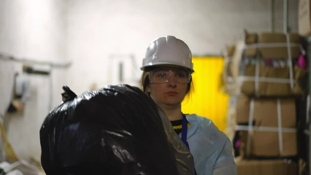 Serious woman with garbage bag walking on recycling station as live camera follows worker. Portrait of focused concentrated Caucasian lady in uniform strolling with trash indoors