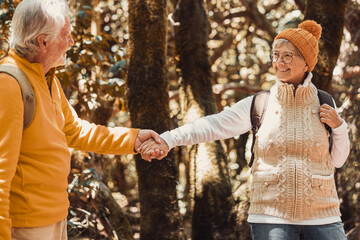 Cheerful caucasian senior couple hand in hand hiking in the forest holding backpacks enjoying freedom, nature and healthy retirement lifestyle