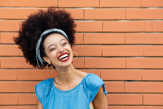 Cheerful young ethnic woman with Afro hair standing near brick wall