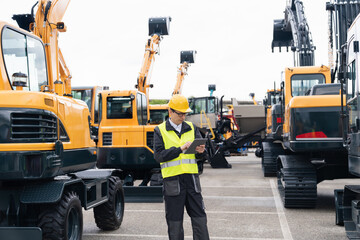 Engineer in a helmet with a digital tablet stands next to construction excavators.