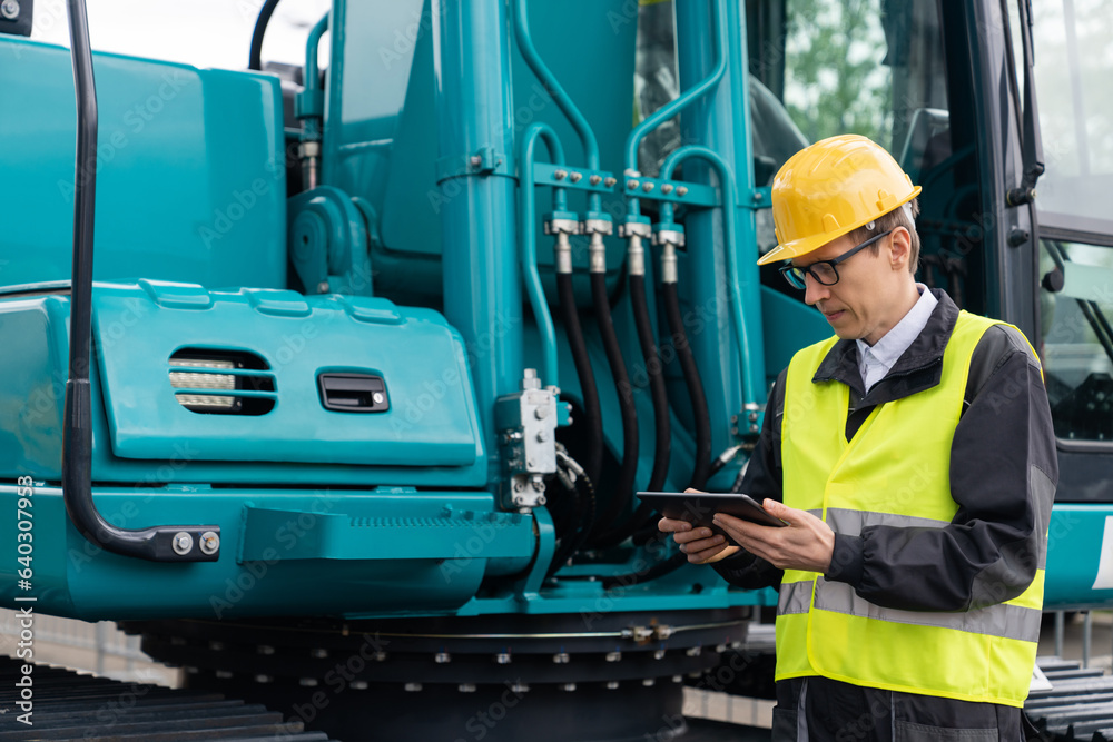 Poster engineer in a helmet with a digital tablet stands next to construction excavators.