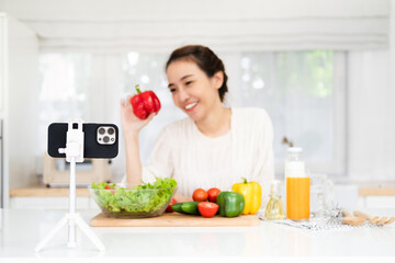 Young cute Asian blogger girl recording video tutorial session of salad cooking lesson at home kitchen. Food blogging or vlogging, social media hobby broadcasting, or online learning course concept