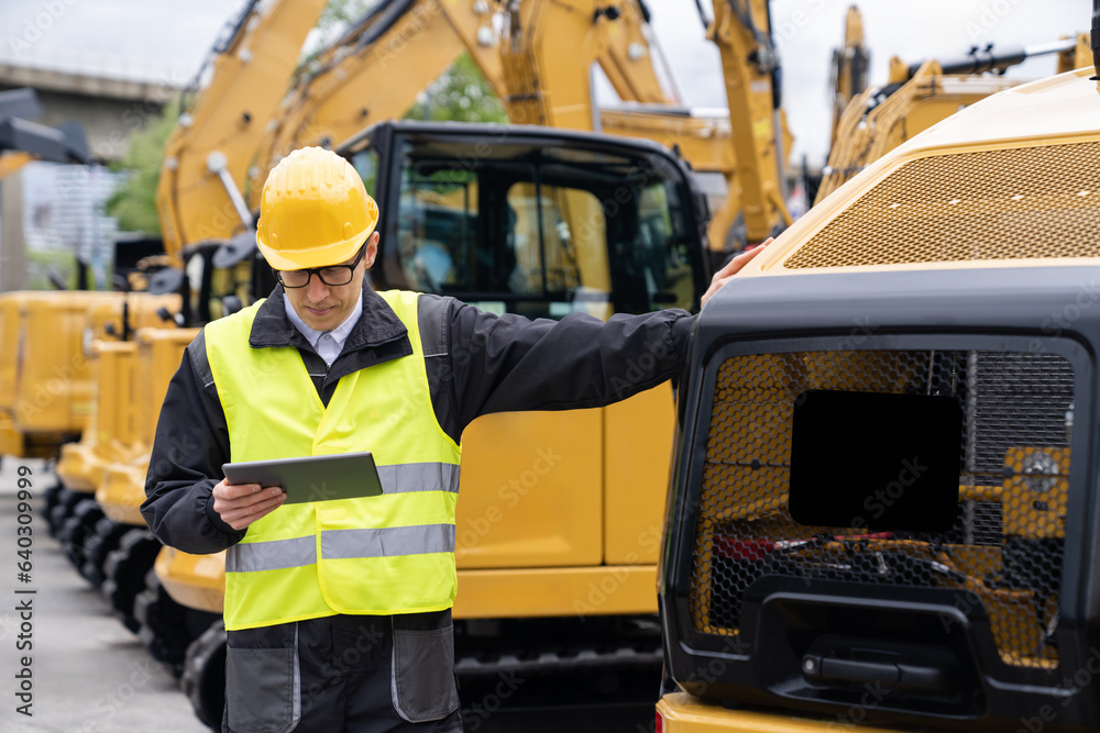 Poster engineer in a helmet with a digital tablet stands next to construction excavators.