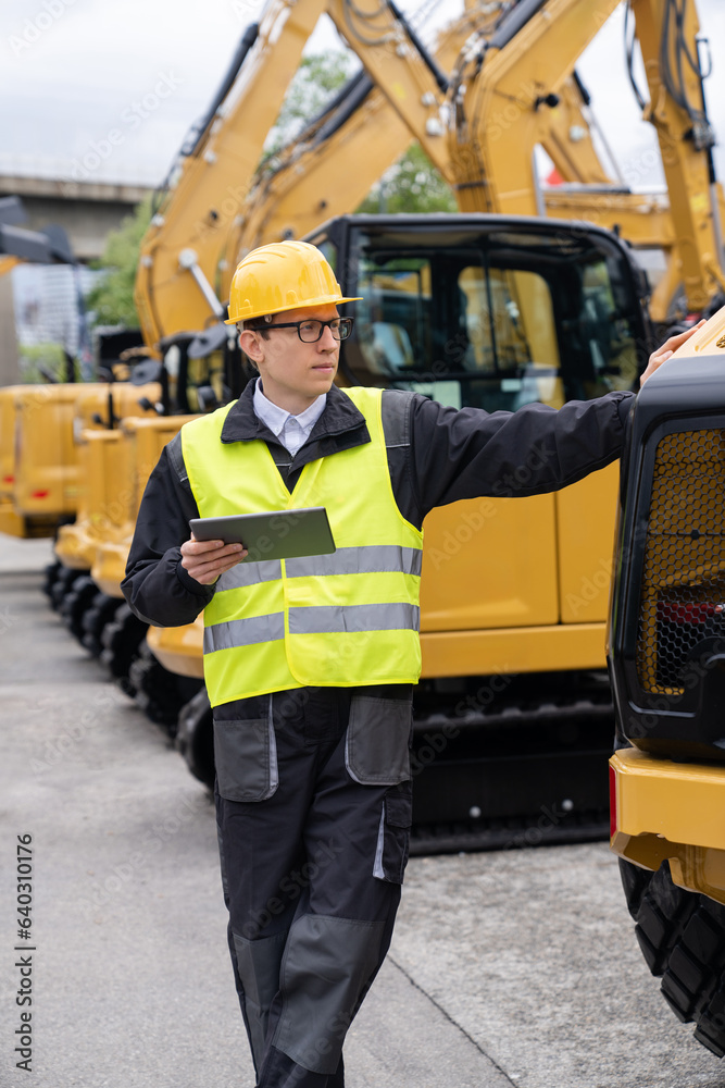 Poster engineer in a helmet with a digital tablet stands next to construction excavators.