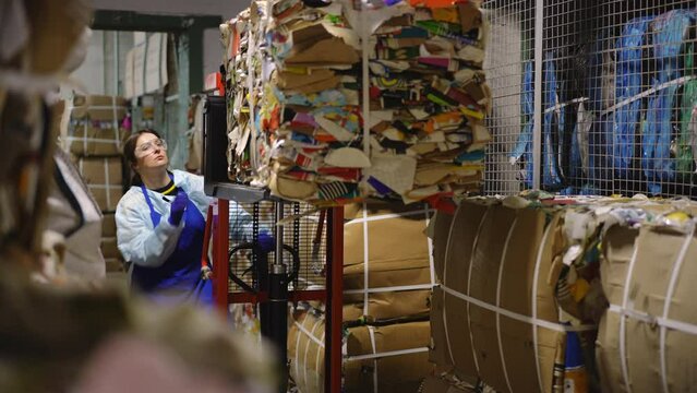 Portrait of focused young woman in uniform lifting packed paper trash with manual forklift in slow motion. Concentrated female Caucasian worker raising garbage for recycling indoors