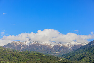 A scenics view of a green mountain valley with snowy rocky mountain summits under a majestic blue sky and some white clouds
