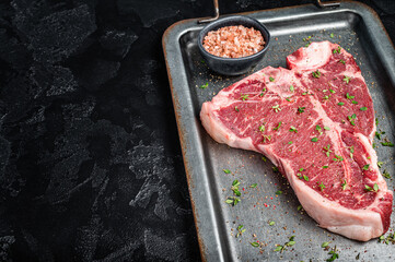 T-bone steak, raw marbled beef meat in a steel kitchen tray with herbs. Black background. Top view. Copy space