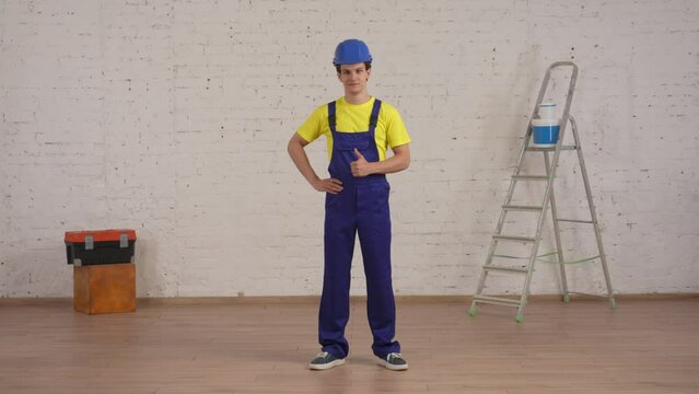 Full length video of a smiling young construction worker standing in the room under renovation with his hand on his hip and giving a thumbs up.