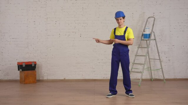 Full length video of a smiling young construction worker standing in the room under renovation pointing to the side of the frame. Place for your advertisement.