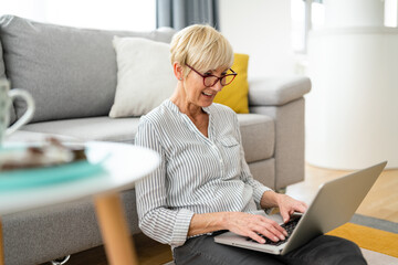 Smiling happy older woman with glasses using laptop for electronic banking, concentrated on paying...