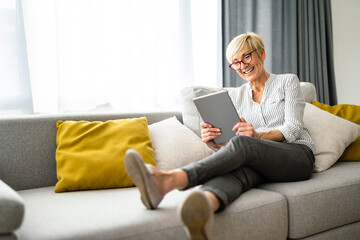 Widely smiling cheerful senior woman reading news on digital tablet while sitting on couch in...
