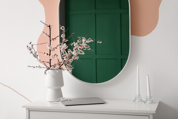 Vase with blooming branches, laptop and candles on commode near mirror in room