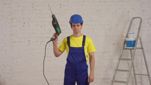 Medium video of a young construction worker standing in the room under renovation, raising the drill, perforator in the air, pointing and giving a thumbs up.