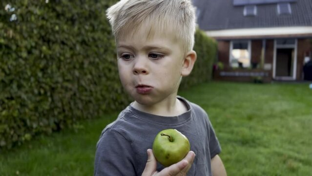 Close up of a cute little toddler boy eating a green apple at home in the garden