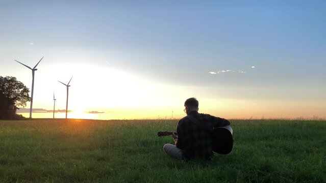 Guitarist musician playing acoustic guitar at sunset sitting in a green field against the background of the evening sky on a farm with windmills in the background