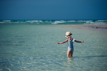 Rear view of a beautiful little girl standing in the water on the beach in a swimsuit and a straw...