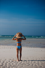 Rear view of a beautiful slim woman standing on the beach in a bikini and a straw hat.