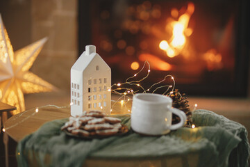 Stylish christmas little house with golden lights, gingerbread cookies and tea cup on table on background of fireplace in festive cozy modern room. Winter hygge. Happy holidays