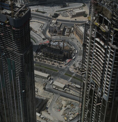 Aerial view looking down onto skyscrapers under construction in Dubai. The unfinished buildings soar high. The background behind them is a mix of ongoing construction and finished projects.