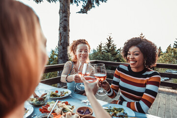 Young and diverse group of female friends enjoying a glass of wine and dinner on a balcony in a cabin house with a scenic view of the mountain and forest below