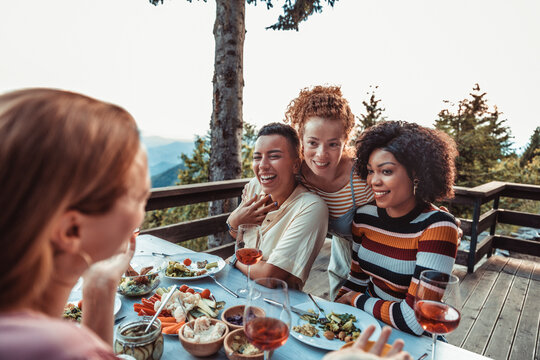 Young and diverse group of female friends enjoying a glass of wine and dinner on a balcony in a cabin house with a scenic view of the mountain and forest below