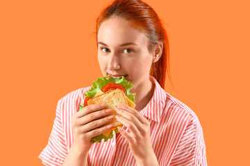 Young redhead woman eating tasty sandwich on orange background, closeup
