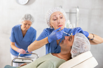Obraz premium Team of cosmetologists provide anti-aging injection services to male patient. Elderly female doctor performs wrinkle smoothing procedure, gives injection of botulinum toxin to man patient in forehead