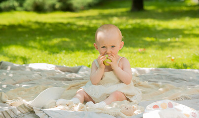 cute baby with an apple in the park.adorable baby sits on a blanket in the parks and eats holding an apple