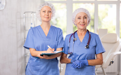 Portrait of doctors with anamnesis questionnaire. Mature woman doctor and female colleague in disposal cap read medical documents, discuss results of blood tests of patient while standing in hospital - obrazy, fototapety, plakaty