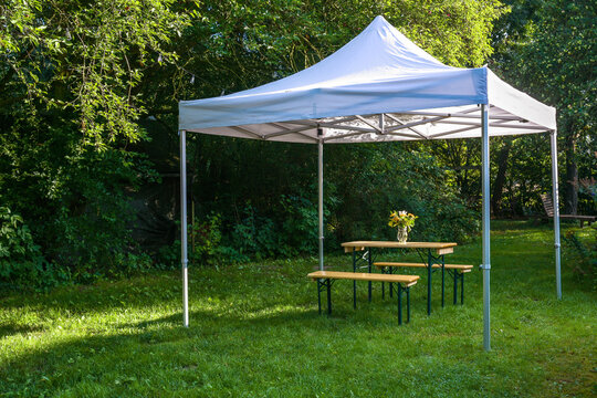 White gazebo or pavilion with a wooden table, benches and a flower bouquet on the meadow in a natural garden, idyllic summer place gives protection from sun and rain, copy space