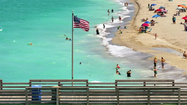 American flag waving over sandy beach near Venice fishing pier in Florida. Tourists enjoying vacation time swimming and tanning. Seaside summer activities