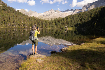 Fototapeta na wymiar Gray-haired man with backpack photographing beautiful landscape with mountain lake and reflection, Austria
