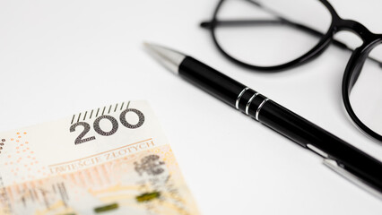 a banknote of Polish zlotys lying on a white background, glasses and a black pen next to it (selective focus)