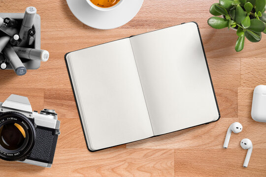 Top view of an opened blank hardcover leather notebook on a wood office desk table with  with pens, cup of coffee, earphone, vintage film camera and succulent plant. High resolution.
