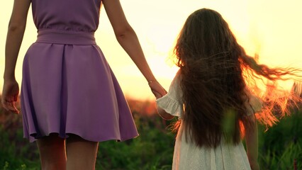 Little daughter with mother join hands walking across park field at sunset light
