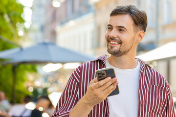Happy smiling brunette Caucasian man using smartphone typing text messages browsing internet social media web app working chatting online outdoor. Guy tourist walking in urban city street. Lifestyles