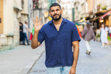 Bearded indian man smiling friendly at camera, waving hands gesturing invitation hello, hi, greeting or goodbye, welcoming with hospitable expression outdoors. Guy walking in urban city town street