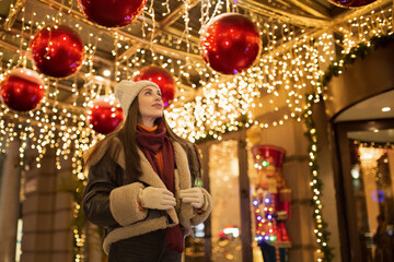 Happy young woman on Christmas night looks up. Bright elegant street and glowing lanterns