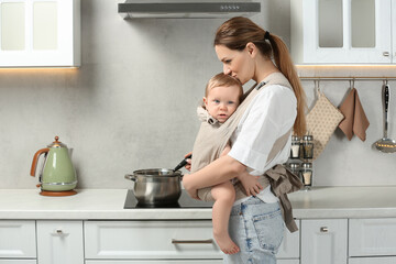 Mother holding her child in sling (baby carrier) while cooking in kitchen