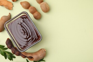 Bowl of tasty tamarind sauce, leaves and fresh pods on light green table, flat lay. Space for text
